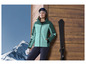Een vrouw in een groene ski-jas met ski's en handschoenen.