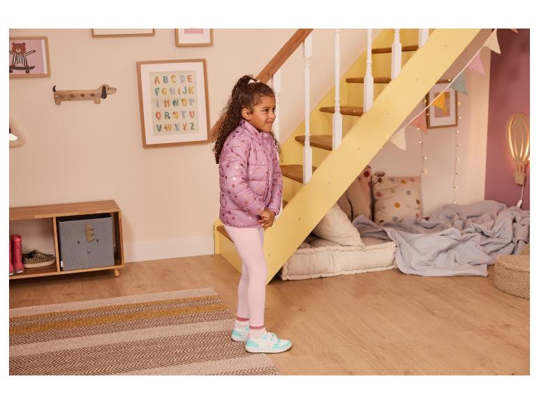 Meisje in een roze pufferjack en lichtroze legging in een speelse kamer.