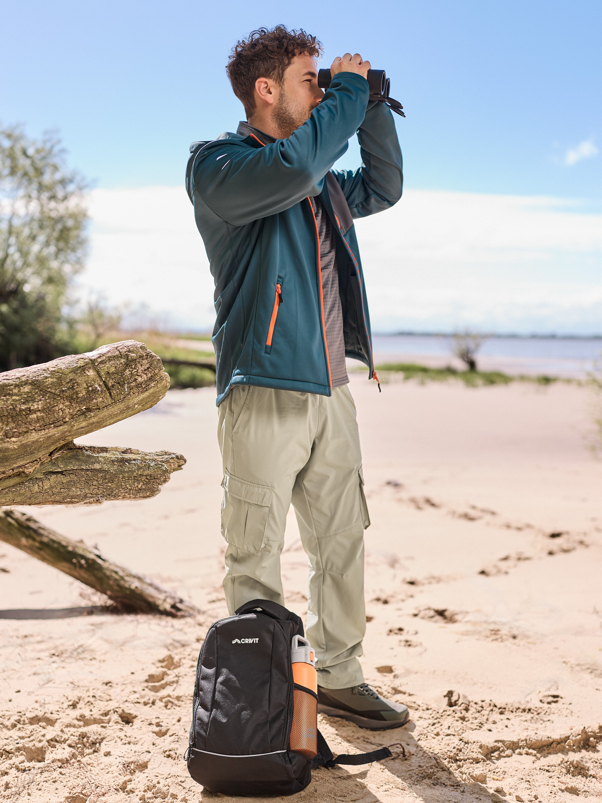 Man in wandeljas en -broek, verrekijker, rugzak en waterfles op het strand