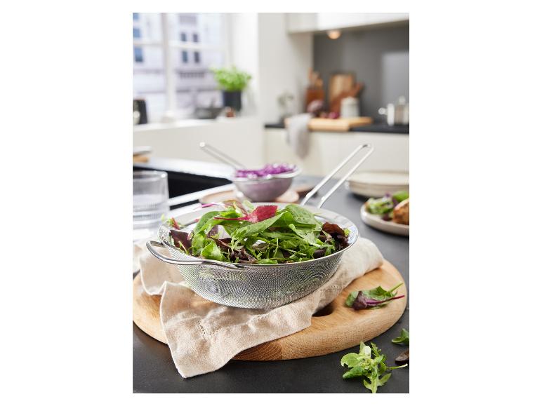Une passoire en métal avec de la salade sur une planche à découper en bois dans la cuisine.
