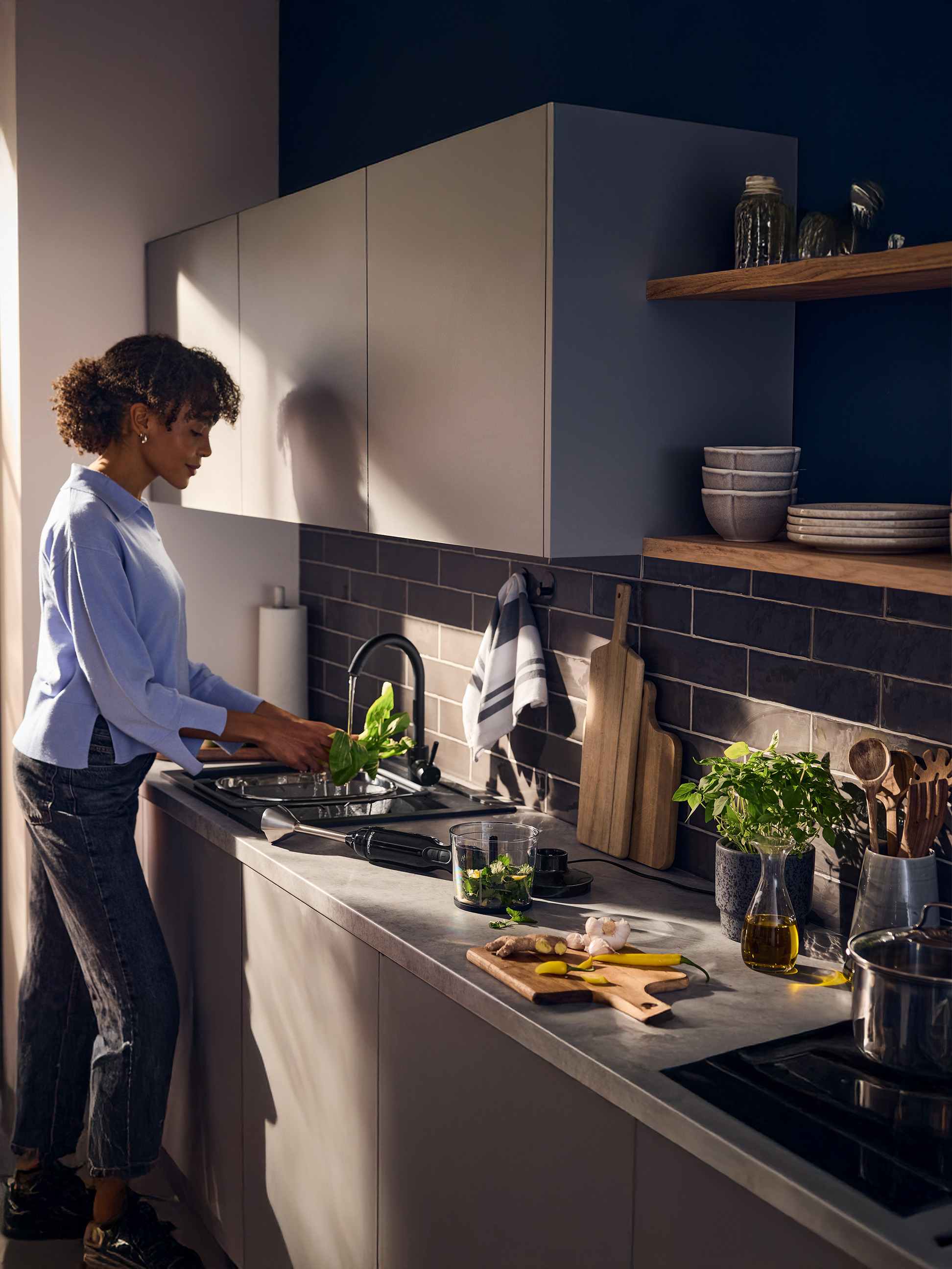 Vrouw wast verse kruiden in de gootsteen, met staafmixer en ingrediënten op het aanrecht.