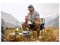 Homme en casquette et t-shirt, tenant une tasse, assis sur une chaise de camping avec une lanterne.
