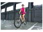 Vrouw in roze vest en helm, rijdend op een elektrische fiets binnen.