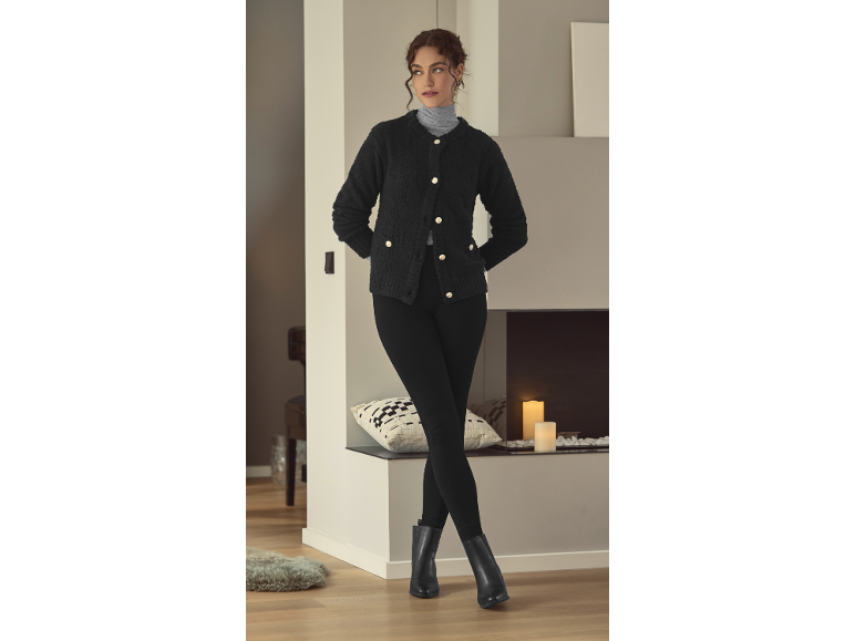Vrouw in zwart vest, coltrui en legging, poserend in een gezellige kamer.