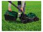 Persoon bevestigt een groen-zwarte grasopvangbak aan een grasmaaier op groen gras.