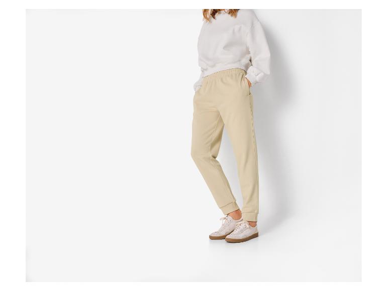 Vrouw draagt beige sweatshirt en beige joggingbroek op witte achtergrond.