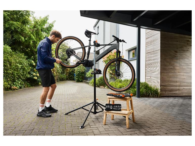 Homme réparant un vélo de montagne gris sur un support de réparation, avec des outils sur un tabouret.