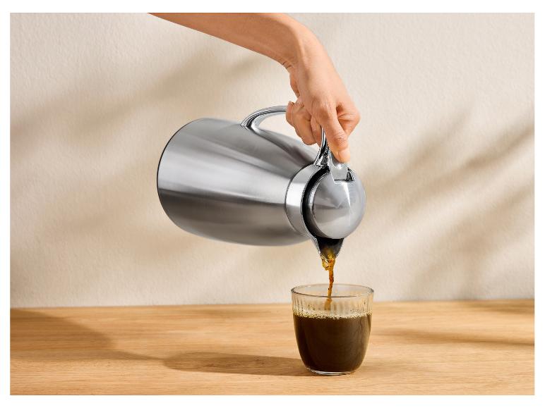 Main versant du café d'un thermos argenté dans un verre sur une table en bois.