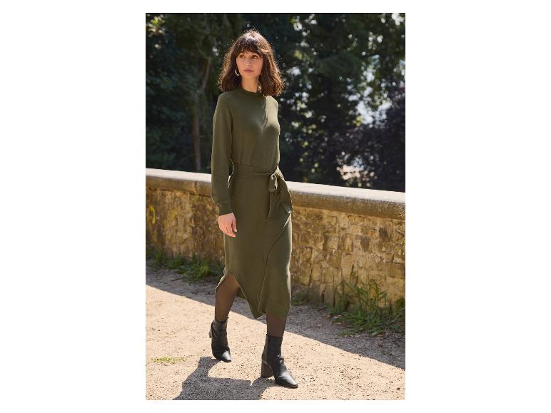 Femme en robe midi vert olive avec des bottes noires marchant sur une allée.
