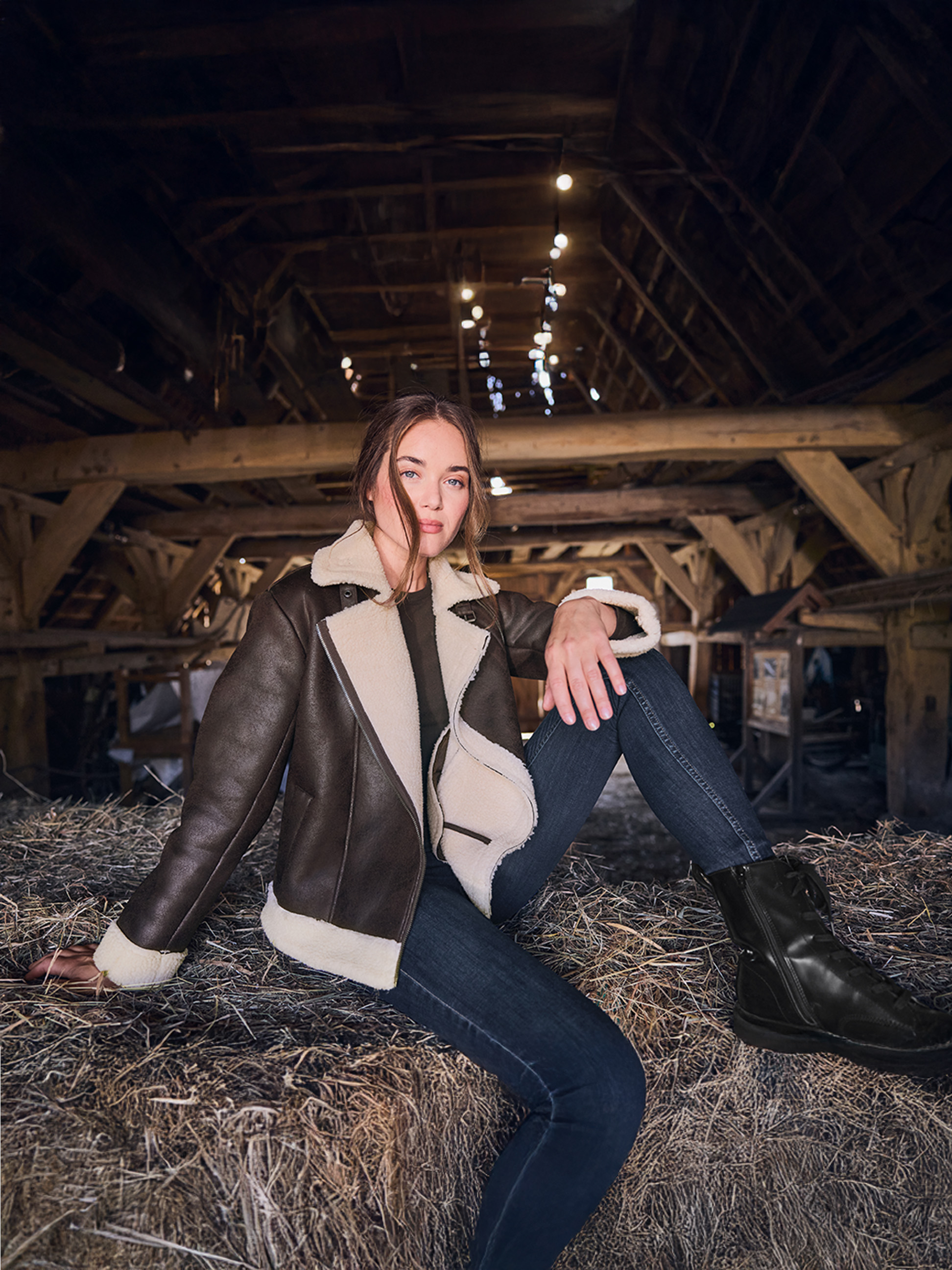Femme en veste shearling marron et jean, assise sur des bottes de foin dans une grange.