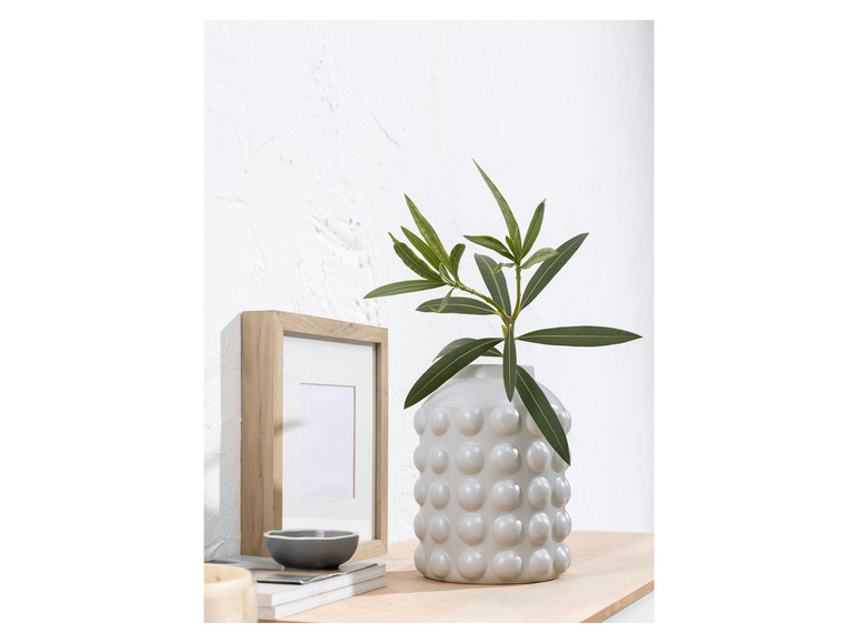 Un vase gris avec des bosses, rempli de branches vertes, à côté d'un cadre en bois.