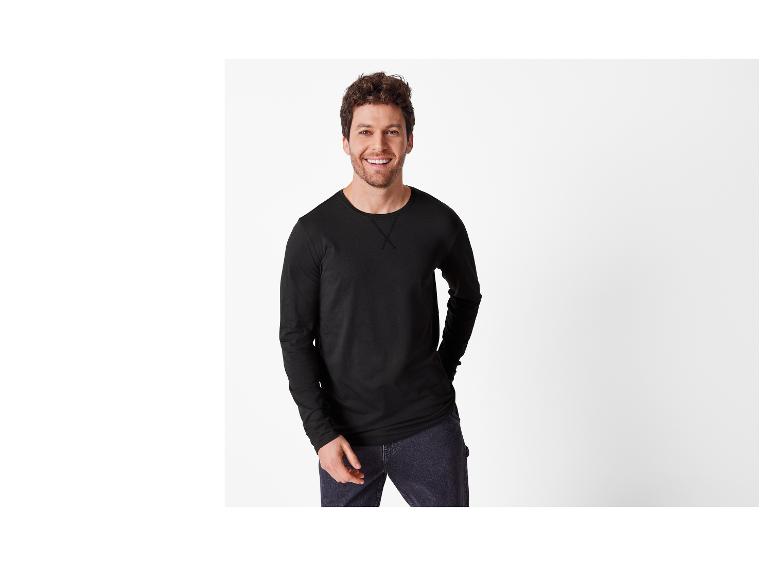 Un homme souriant en T-shirt noir à manches longues et jean foncé.