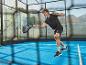 Man in sportkleding speelt padel met racket en bal op een blauwe baan.