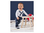Een baby in een wit shirt met een blauwe strik en een blauwe broek met bootjes staat bij een houten krukje.