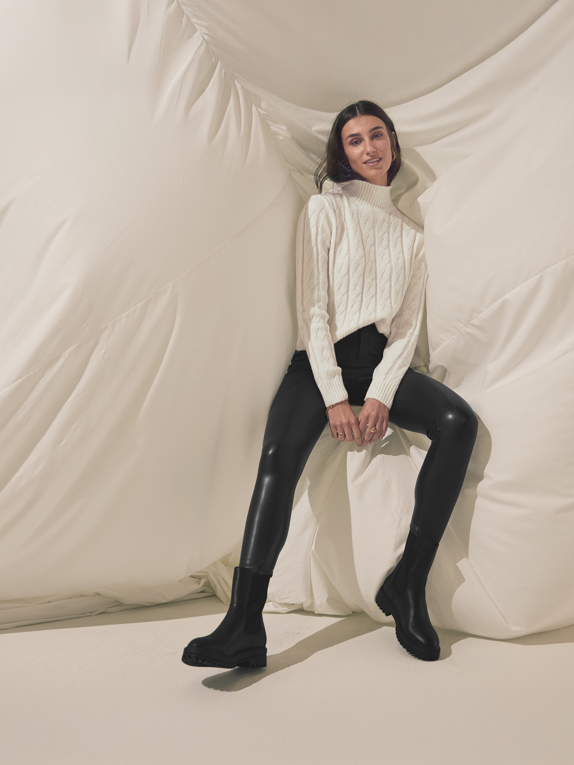 Vrouw in witte kabeltrui, zwarte imitatie leren legging en zwarte laarzen.