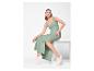 Femme en robe longue verte et baskets blanches, assise sur un piédestal blanc.