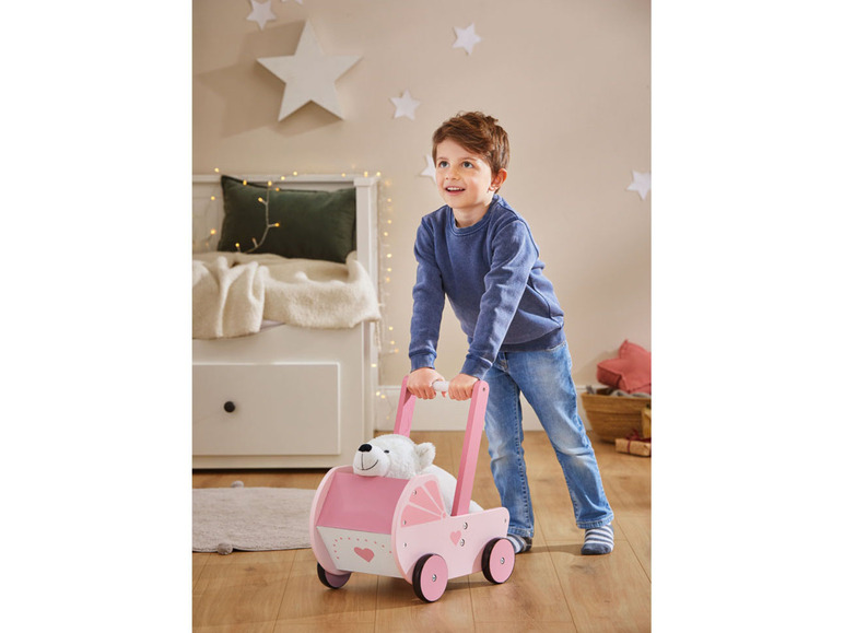 Un garçon pousse un chariot en bois rose avec un ours en peluche.
