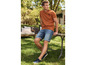 Man in een oranje T-shirt en blauwe denim shorts, zittend op een houten bankje in de tuin.