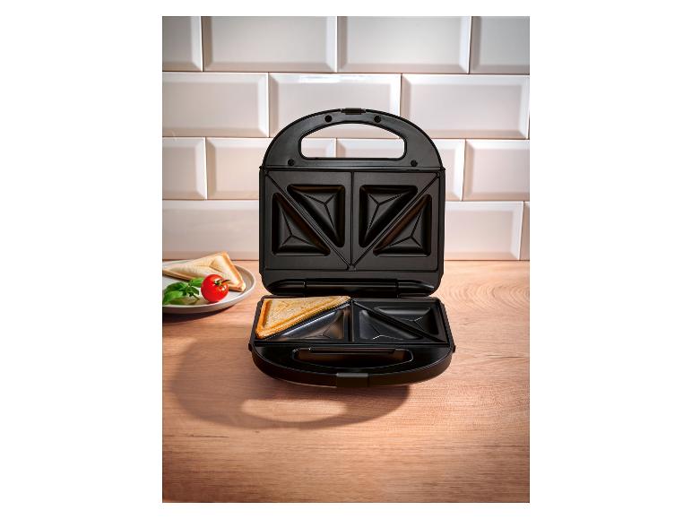 Open tosti-ijzer met tosti en een bord met tomaat en basilicum op een houten aanrecht.