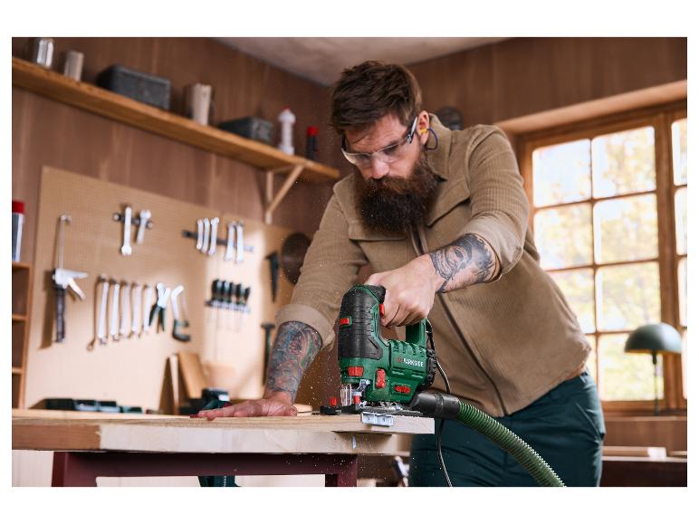 Een bebaarde man gebruikt een elektrische zaag om een houten plank in een werkplaats te zagen.
