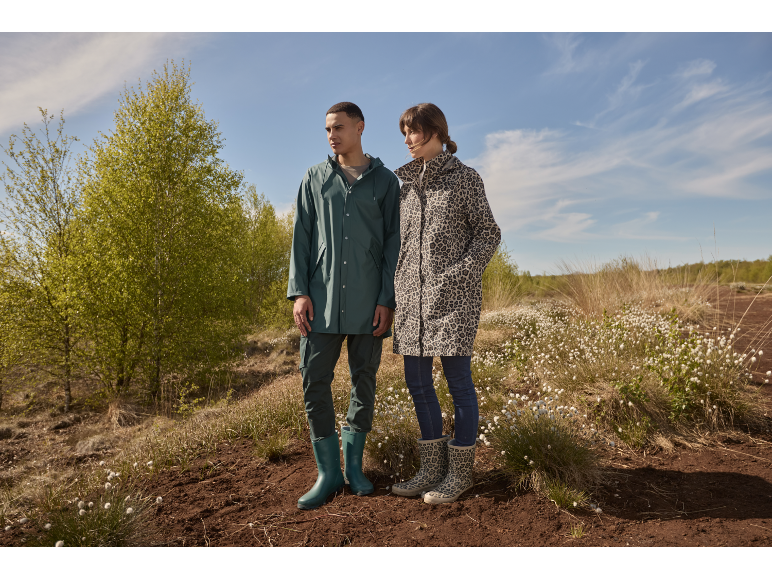 Man en vrouw in regenjassen en laarzen staan in een veld met witte bloemen.