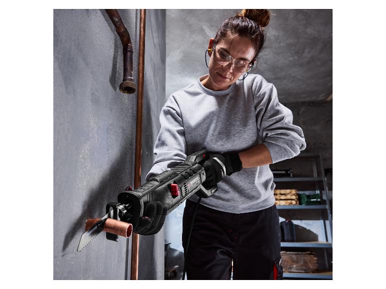 Femme coupant un tuyau en cuivre avec une scie sabre électrique, portant des lunettes de sécurité et des gants.