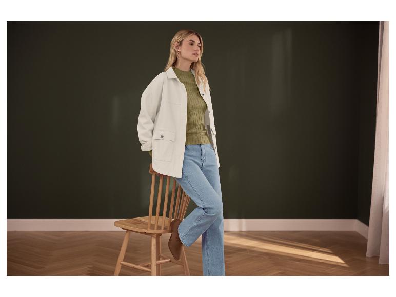Vrouw in groene trui, wit jack en blauwe jeans, leunend tegen een houten stoel.