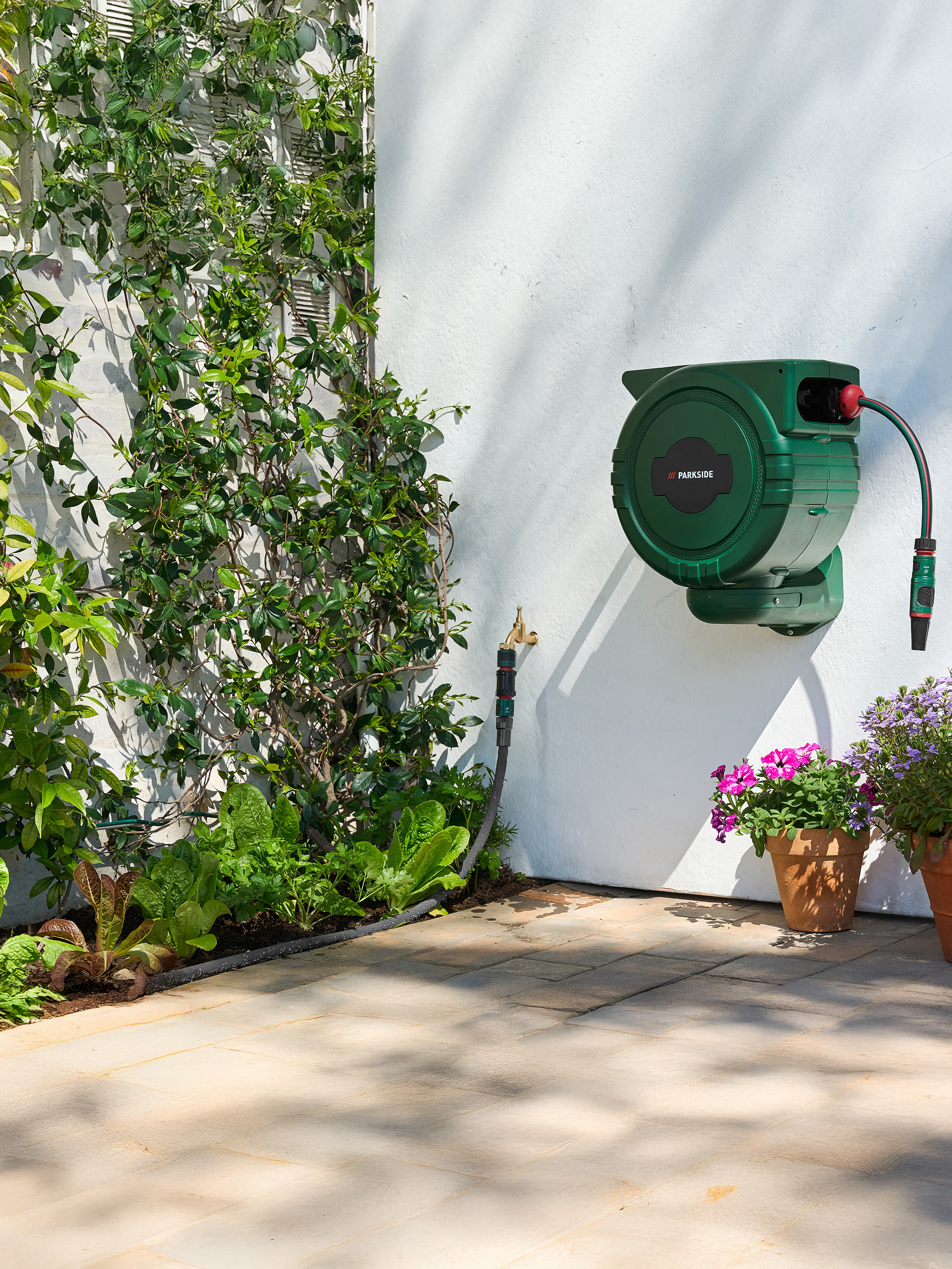 Groene PARKSIDE tuinslanghaspel aan een witte muur met klimplanten en een moestuin.