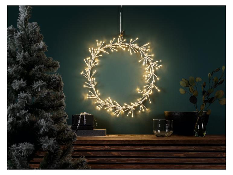 Lichtgevende kerstkrans aan een groene muur, met een kerstboom en decoraties.