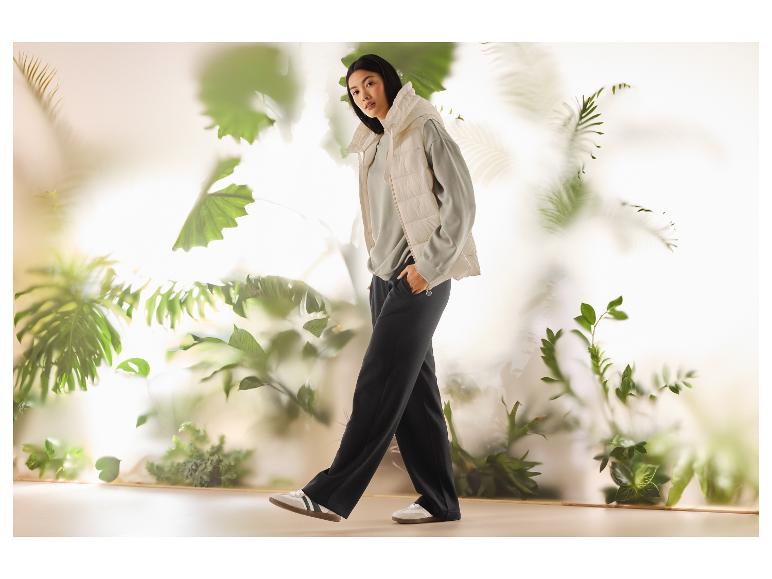 Femme en gilet matelassé beige, sweat-shirt vert et pantalon noir, marchant parmi les plantes.