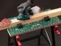 Een gehandschoende hand gebruikt een elektrische schuurmachine op een werkbank met een houten plank.