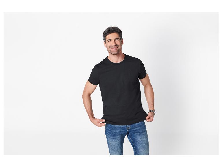 Man in zwart T-shirt en blauwe jeans, lachend tegen een witte achtergrond.
