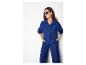 Femme en chemise bleue brodée et pantalon assorti, souriante les mains dans les poches.