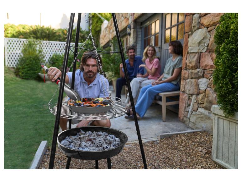 Man grilt zeevruchtenpaella op een driepootbarbecue met vrienden op de achtergrond.