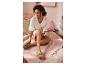 Femme en robe blanche et sandales vertes assise sur une couverture rose avec des coussins.