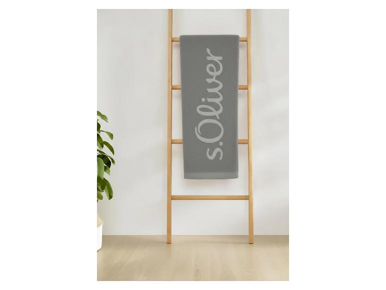 Serviette grise S.Oliver avec logo, suspendue à une échelle en bois, à côté d'une plante.