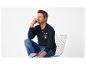 Homme souriant en polo à manches longues bleu marine avec motif de voile et jean, assis sur une chaise.
