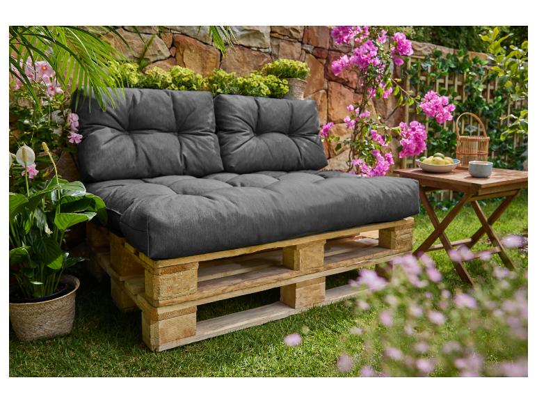 Buitenbank van pallets met donkergrijze kussens, omringd door weelderig groen en roze bloemen.