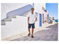 Man in een wit polo shirt en blauwe shorts, loopt op straat met witte huizen.
