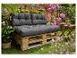 Buitenbank van pallets met donkergrijze kussens, omringd door weelderig groen en roze bloemen.
