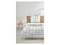 Wit beddengoed met grijs ruitpatroon op bed met houten hoofdbord, nachtkastje met lamp ernaast.