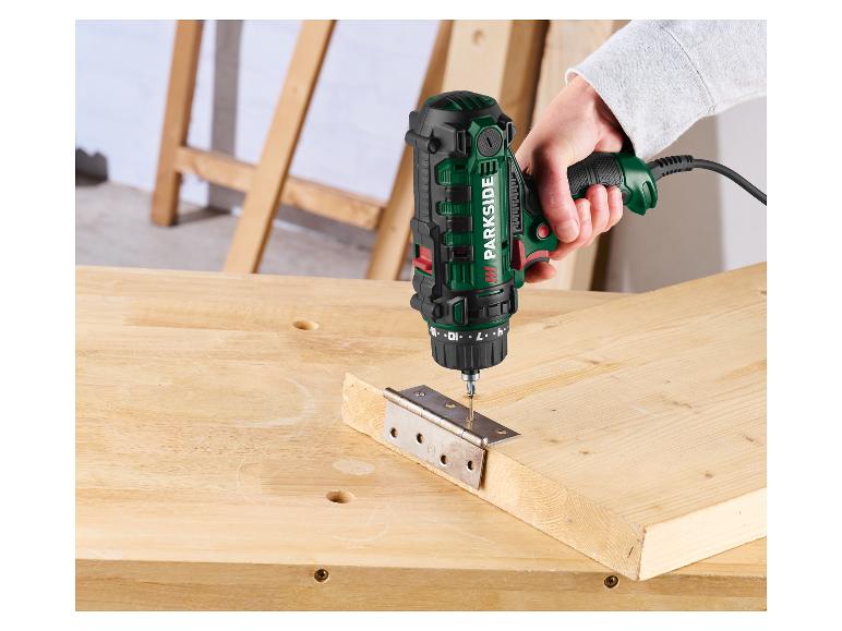 Perceuse/visseuse électrique verte et noire fixant une charnière métallique sur une planche en bois.