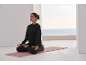 Femme en tenue de sport noire médite sur un tapis de yoga rose avec vue sur la mer.