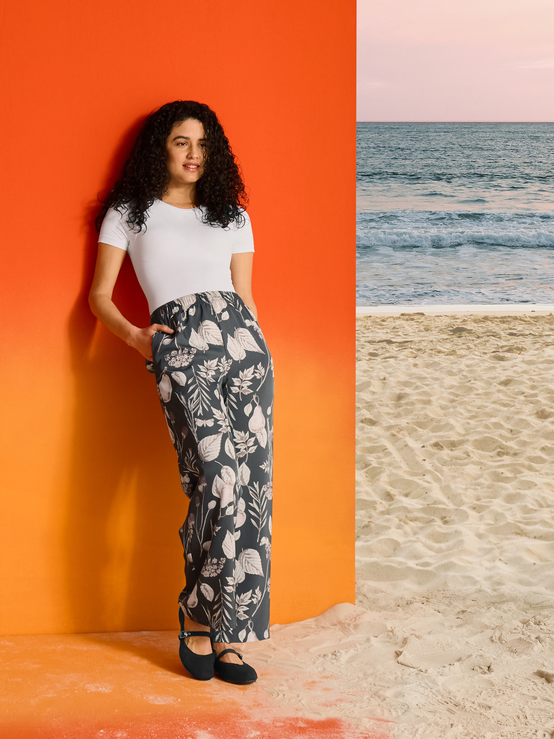 Femme en pantalon à motifs floraux et t-shirt blanc, sur fond orange et plage.