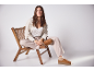Vrouw zit op houten stoel, draagt beige broek, licht overhemd en bruine schoenen.