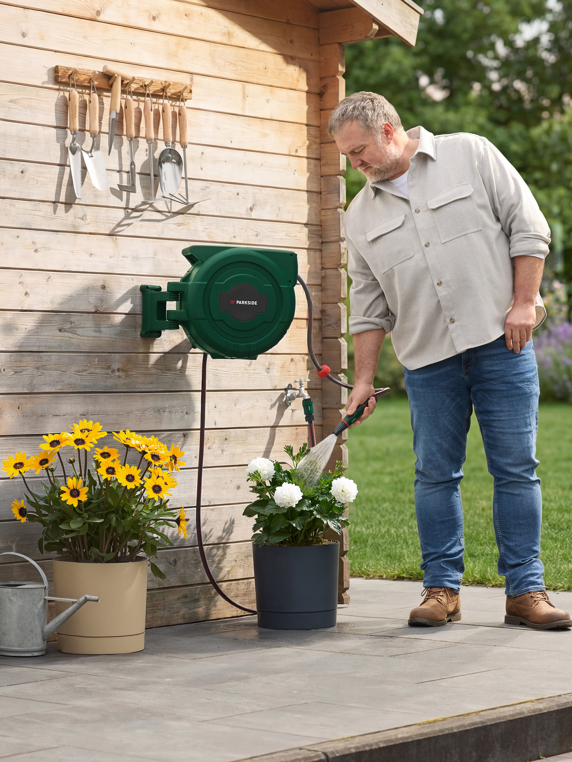 Man besproeit bloemen met een wandslanghaspel en hangend tuingereedschap.