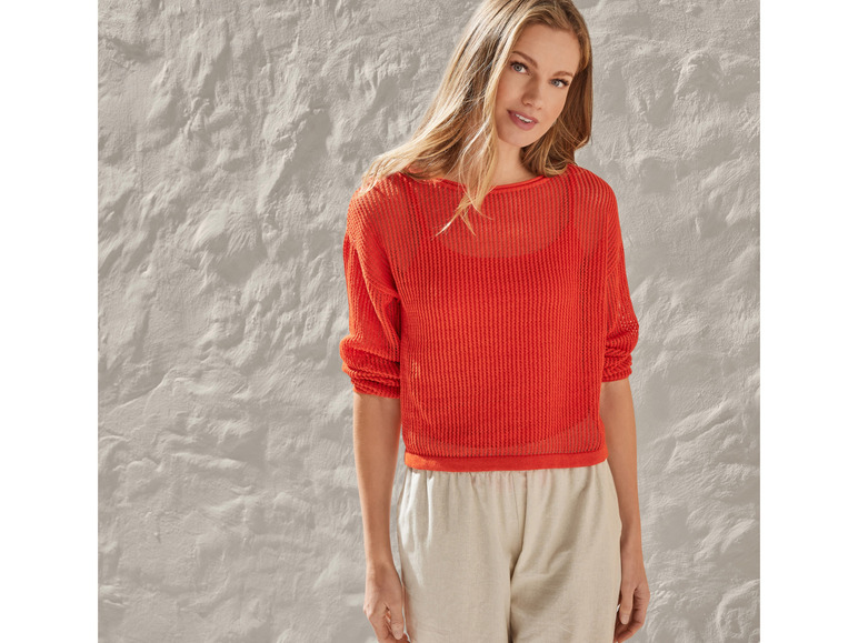 Een vrouw draagt een oranje gebreide trui en een beige broek.