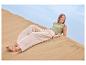 Femme en top vert et pantalon beige sourit allongée sur une dune de sable.