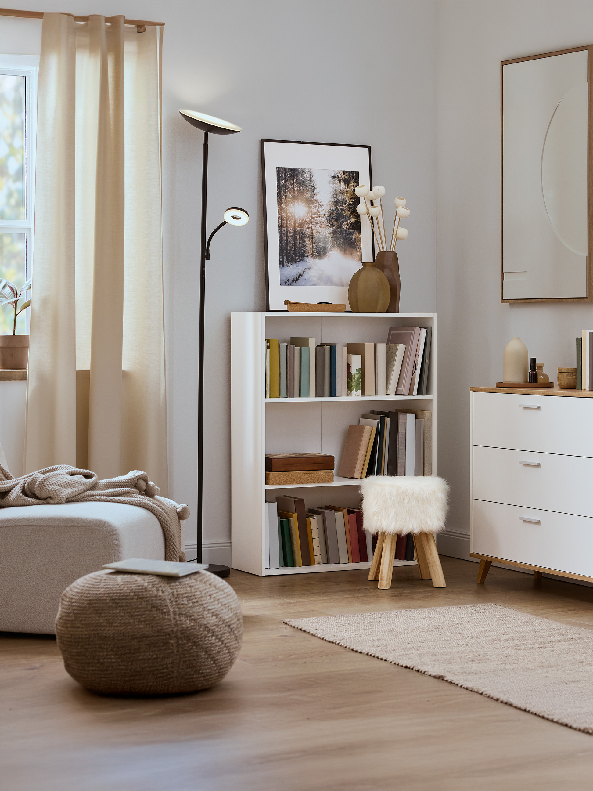 Salon lumineux avec une bibliothèque blanche, un pouf tricoté et un lampadaire moderne.