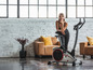 Une femme pose à côté d'un vélo d'appartement noir dans un salon moderne.
